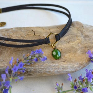 May include: Black choker necklace with a green oval pendant. The necklace features a double-strand black cord and a gold-colored clasp. Lavender sprigs frame the necklace on a wooden surface, creating a natural aesthetic.