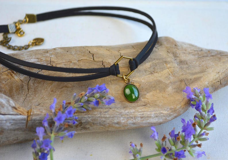 May include: Black choker necklace with a green oval pendant. The necklace features a double-strand black cord and a gold-colored clasp. Lavender sprigs frame the necklace on a wooden surface, creating a natural aesthetic.