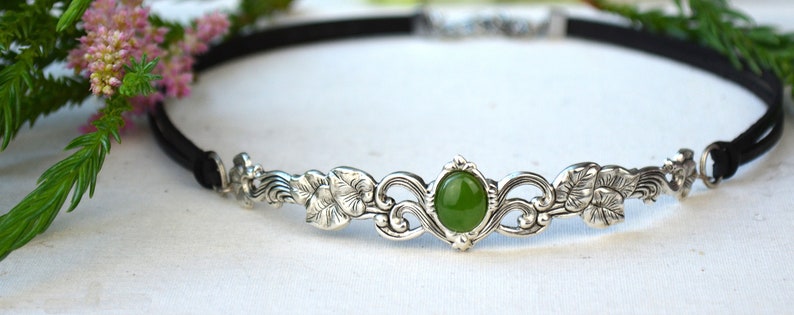 May include: A silver headband with a green gemstone and floral design. The headband is made of a black leather band.