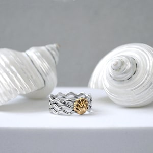 May include: A silver ring with a wave design and a gold seashell accent. Two large white seashells are in the background. The ring is on a white surface, with a soft gray background.
