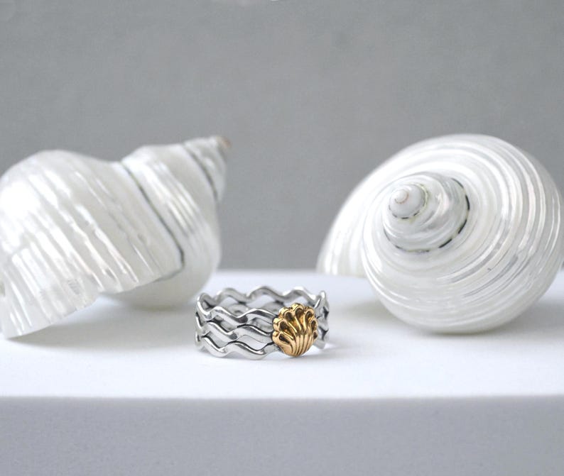 May include: A silver ring with a wave design and a gold seashell accent. Two large white seashells are in the background. The ring is on a white surface, with a soft gray background.