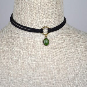 May include: Black choker necklace with a gold-colored hexagonal connector and a green oval pendant. The necklace is displayed on a mannequin with a neutral-colored fabric.
