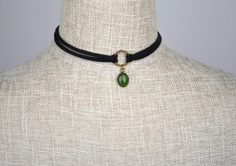 May include: Black choker necklace with a gold-colored hexagonal connector and a green oval pendant. The necklace is displayed on a mannequin with a neutral-colored fabric.