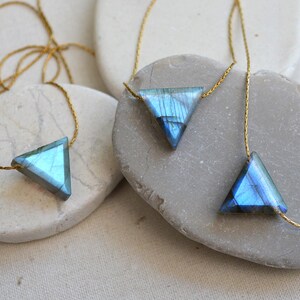 May include: Three necklaces with triangular pendants made of blue and gray iridescent stone. The pendants are suspended from delicate gold-colored chains. The necklaces are displayed on smooth, light-colored stones.