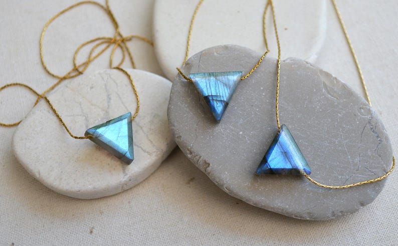 May include: Three necklaces with triangular pendants made of blue and gray iridescent stone. The pendants are suspended from delicate gold-colored chains. The necklaces are displayed on smooth, light-colored stones.
