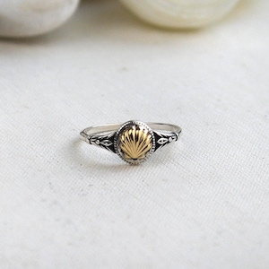 May include: A silver ring with a gold-colored oval centerpiece. The centerpiece has a textured, fan-like design. The ring band features decorative detailing on the sides. The ring is set against a white background.