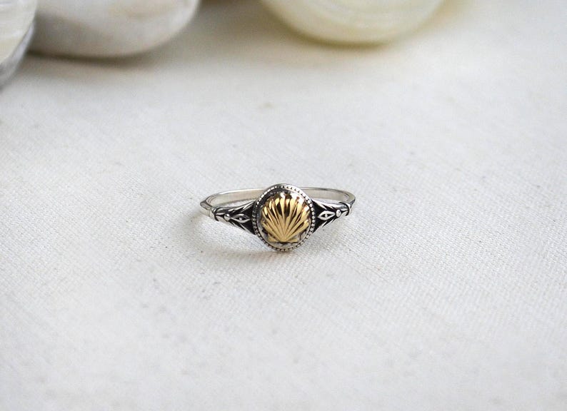 May include: A silver ring with a gold-colored oval centerpiece. The centerpiece has a textured, fan-like design. The ring band features decorative detailing on the sides. The ring is set against a white background.