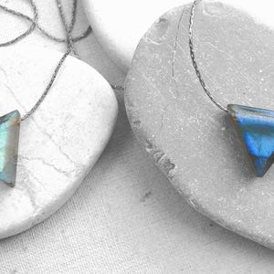 May include: Two triangle-shaped pendant necklaces. One pendant is a light blue-green color, and the other is a deep blue. Both pendants are on a thin, silver-colored chain. The necklaces are displayed on gray stones.