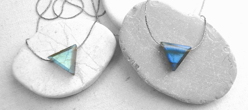 May include: Two triangle-shaped pendant necklaces. One pendant is a light blue-green color, and the other is a deep blue. Both pendants are on a thin, silver-colored chain. The necklaces are displayed on gray stones.