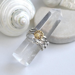 May include: A silver ring with a gold seashell design, resting on a clear crystal. The ring has a wavy band and a detailed gold seashell centerpiece. A white seashell is in the background.