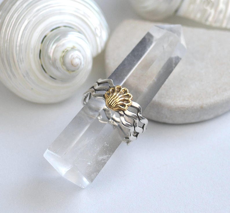 May include: A silver ring with a gold seashell design, resting on a clear crystal. The ring has a wavy band and a detailed gold seashell centerpiece. A white seashell is in the background.