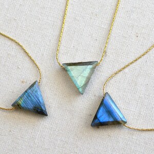 May include: Three triangle-shaped labradorite stone pendants on delicate gold chains. The stones display iridescent blue and green hues. The necklaces are displayed on a neutral, textured surface, showcasing the jewelry's design.
