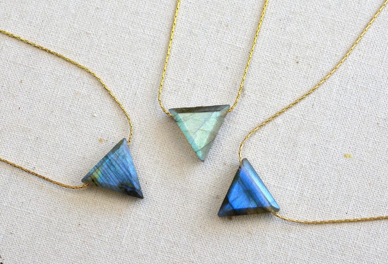 May include: Three triangle-shaped labradorite stone pendants on delicate gold chains. The stones display iridescent blue and green hues. The necklaces are displayed on a neutral, textured surface, showcasing the jewelry's design.