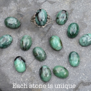 May include: A collection of 11 oval-shaped green gemstones with black inclusions. The stones are arranged on a gray surface. Each stone is unique. "Each stone is unique" is written in black text at the bottom of the image.