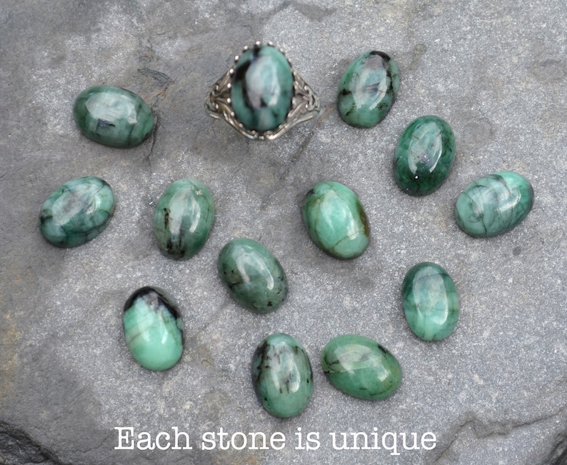 May include: A collection of 11 oval-shaped green gemstones with black inclusions. The stones are arranged on a gray surface. Each stone is unique. "Each stone is unique" is written in black text at the bottom of the image.