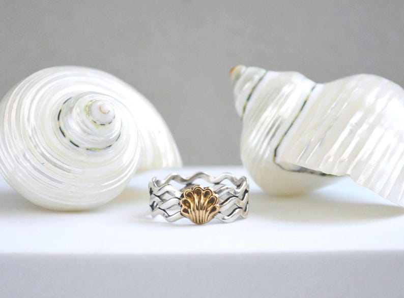 May include: A silver ring with a wavy design and a gold seashell in the center. The ring is displayed in front of two large white seashells on a white surface. The ring's design is reminiscent of ocean waves.