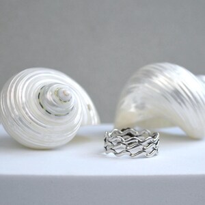 May include: A silver ring with a wave design sits in front of two white seashells. The ring has a repeating pattern of curved lines, creating a wavy appearance. The seashells have a spiral shape and a pearlescent sheen.