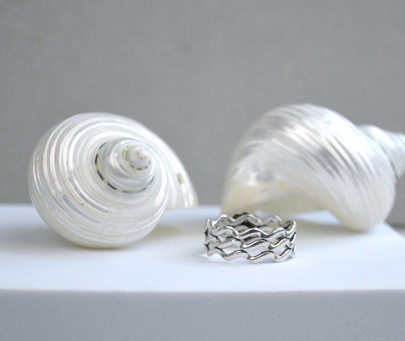 May include: A silver ring with a wave design sits in front of two white seashells. The ring has a repeating pattern of curved lines, creating a wavy appearance. The seashells have a spiral shape and a pearlescent sheen.
