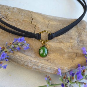 May include: A black choker necklace with a green oval pendant and gold-colored accents. The necklace is displayed on a piece of driftwood, with sprigs of purple lavender flowers on either side.