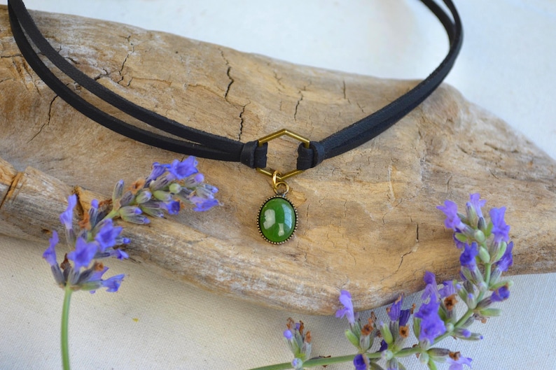 May include: A black choker necklace with a green oval pendant and gold-colored accents. The necklace is displayed on a piece of driftwood, with sprigs of purple lavender flowers on either side.