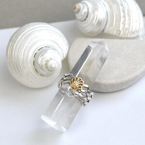 May include: A silver ring with a gold seashell design, resting on a clear crystal. Two white seashells are in the background. The ring has a wavy band and a gold seashell in the center.