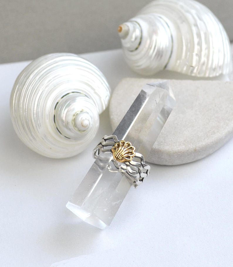 May include: A silver ring with a gold seashell design, resting on a clear crystal. Two white seashells are in the background. The ring has a wavy band and a gold seashell in the center.