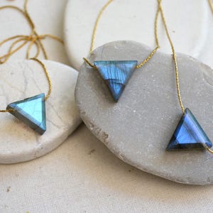 May include: Three triangle-shaped necklaces with iridescent blue and green gemstones. Each pendant is suspended from a delicate gold chain. The necklaces are displayed on smooth, light-colored stones, creating a minimalist aesthetic.