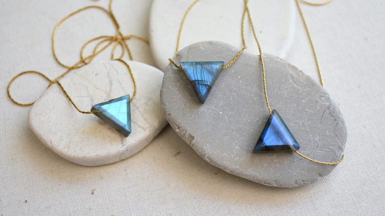 May include: Three triangle-shaped necklaces with iridescent blue and green gemstones. Each pendant is suspended from a delicate gold chain. The necklaces are displayed on smooth, light-colored stones, creating a minimalist aesthetic.