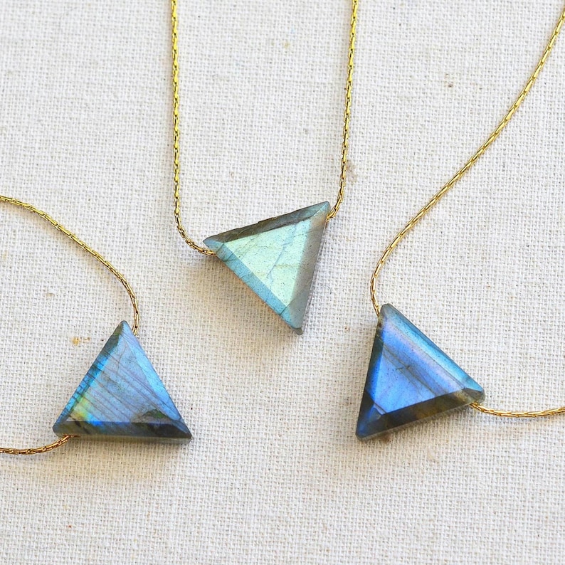 May include: Three triangle-shaped labradorite necklaces with gold-colored chains. The gemstones display iridescent blue, green, and gray hues. The necklaces are displayed on a white, textured surface, showcasing the jewelry's unique design and color variations.