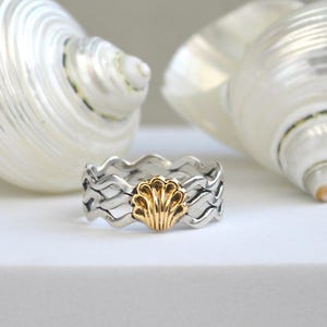 May include: A silver ring with a wavy design and a gold seashell in the center. The ring is set against a white background with two large, white seashells. The ring's design evokes a coastal theme.