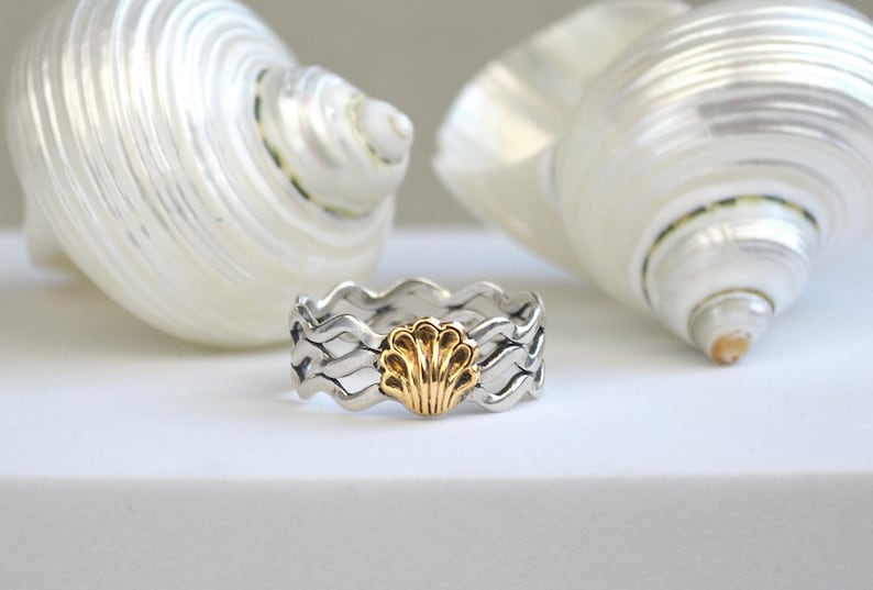 May include: A silver ring with a wavy design and a gold seashell in the center. The ring is set against a white background with two large, white seashells. The ring's design evokes a coastal theme.
