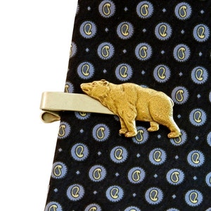 May include: A gold-toned tie clip featuring a bear design. The clip is attached to a dark blue patterned tie.