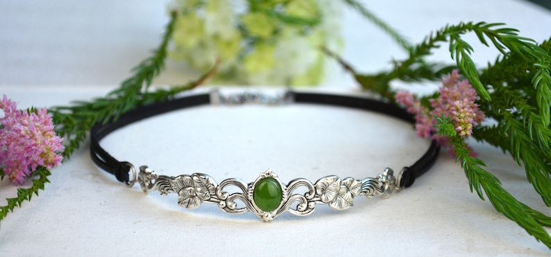 May include: A silver headband with a green gemstone and floral design. The headband is made of a black elastic band.