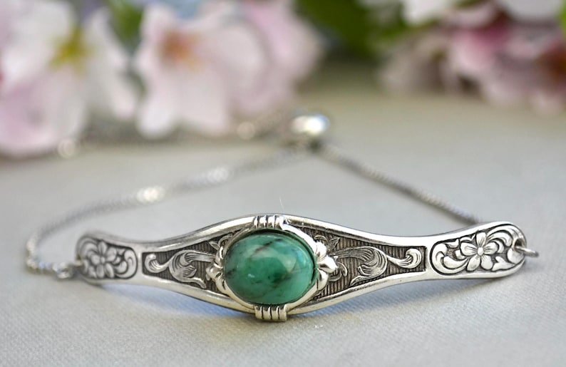 May include: A silver bracelet with an ornate design and a green gemstone. The bracelet has a sliding clasp.