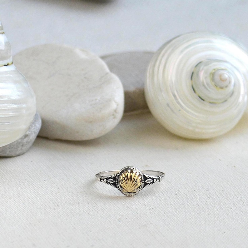 May include: A silver ring with a gold seashell design. The ring has an oval-shaped gold center with a detailed seashell pattern, framed by a silver band with decorative accents. The ring is displayed on a white surface.