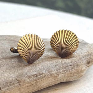 May include: A pair of brass-toned metal cufflinks shaped like fan-shaped seashells. Each cufflink features a detailed, radiating ribbed texture across the surface, mimicking the natural ridges of a scallop shell, with a dark antique finish.