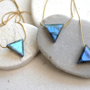 May include: Three triangle-shaped necklaces with iridescent blue and green gemstones on gold chains. The pendants are displayed on smooth, light-colored stones, creating a minimalist and elegant aesthetic. The necklaces are perfect for everyday wear.