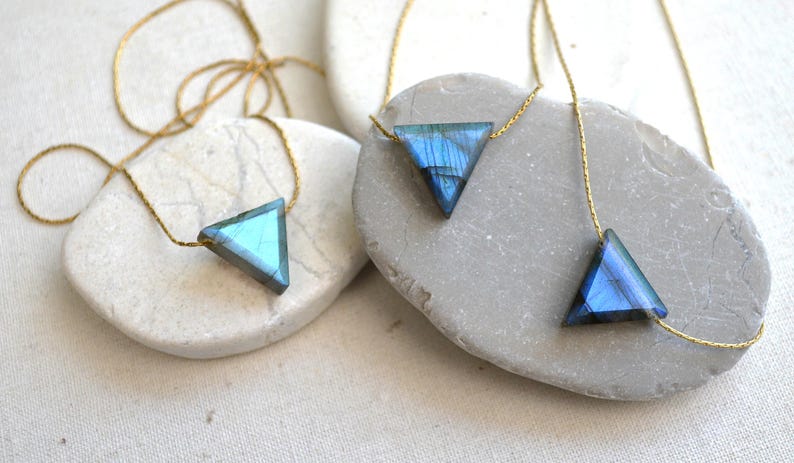 May include: Three triangle-shaped necklaces with iridescent blue and green gemstones on gold chains. The pendants are displayed on smooth, light-colored stones, creating a minimalist and elegant aesthetic. The necklaces are perfect for everyday wear.