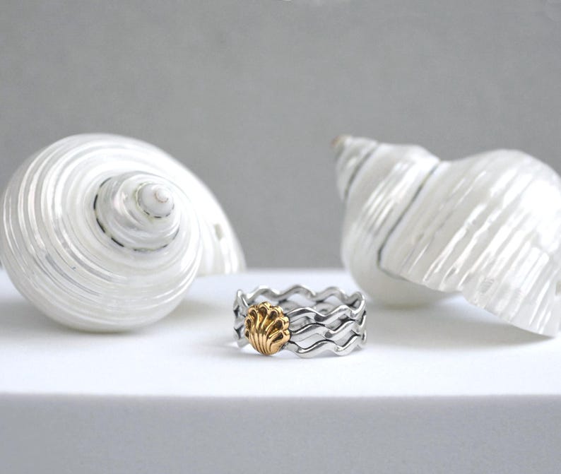 May include: A silver ring with a wavy design and a gold seashell accent. Two large white seashells are in the background. The ring is on a white surface.
