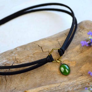 May include: A black choker necklace with a green oval pendant and gold-colored accents. The necklace is made of two thin black cords and rests on a piece of driftwood. Purple flowers are in the background.