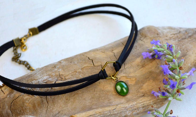May include: A black choker necklace with a green oval pendant and gold-colored accents. The necklace is made of two thin black cords and rests on a piece of driftwood. Purple flowers are in the background.