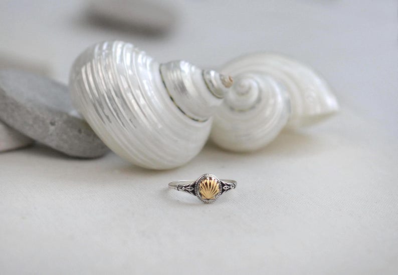 May include: A silver ring with a gold seashell design is displayed in front of a white seashell. The ring has a small, oval-shaped gold center with intricate silver detailing. The background features a large, white seashell and a few gray stones.