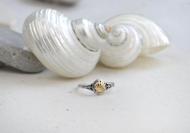 May include: A silver ring with a gold seashell design. The ring is set against a white background with two large, white seashells. The ring's band has intricate detailing.