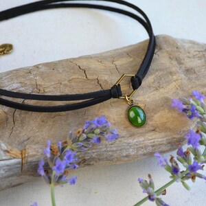 May include: Black choker necklace with a green oval pendant. The necklace features a double-strand black cord and a gold-colored clasp. The pendant is a vibrant green color. The necklace is displayed on a piece of driftwood, with sprigs of purple flowers.