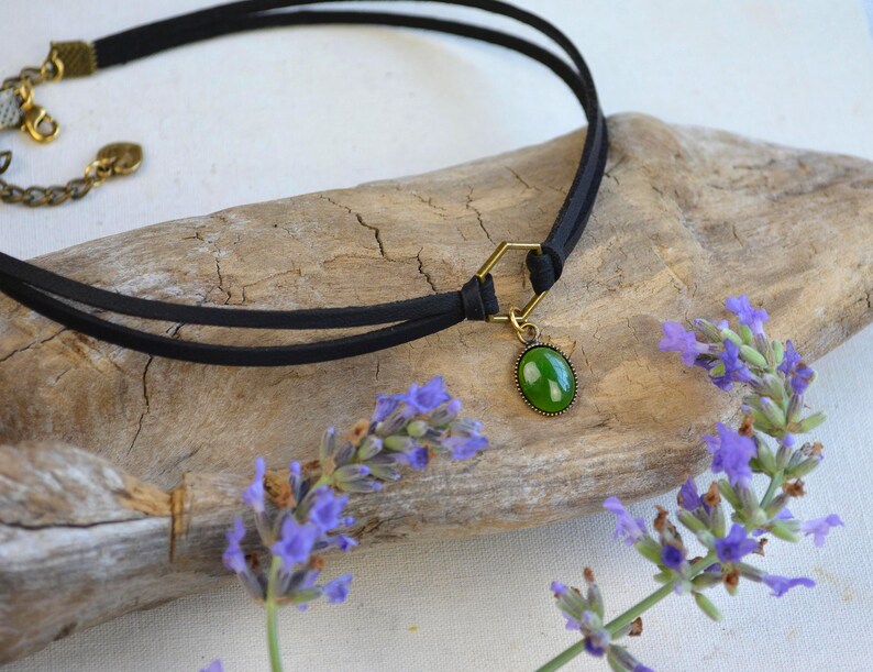May include: Black choker necklace with a green oval pendant. The necklace features a double-strand black cord and a gold-colored clasp. The pendant is a vibrant green color. The necklace is displayed on a piece of driftwood, with sprigs of purple flowers.