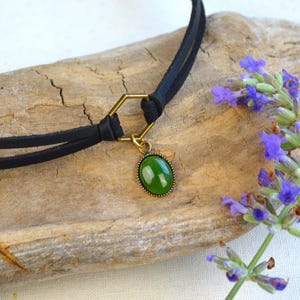 May include: A black choker necklace with a gold-colored hexagonal connector and a green oval pendant. The pendant is set in a gold-colored frame. The necklace is displayed on a piece of driftwood, with sprigs of purple flowers.