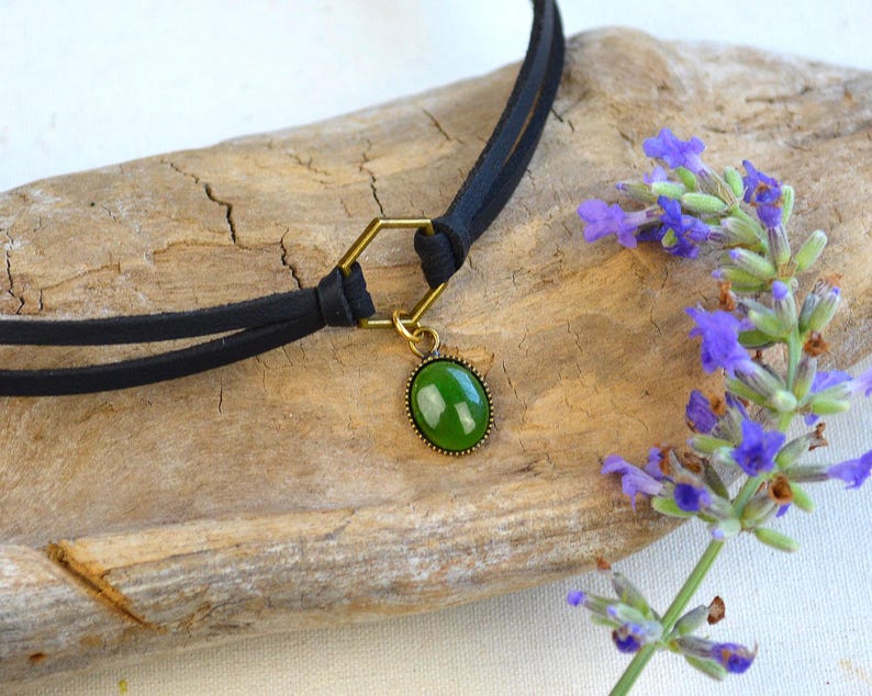 May include: A black choker necklace with a gold-colored hexagonal connector and a green oval pendant. The pendant is set in a gold-colored frame. The necklace is displayed on a piece of driftwood, with sprigs of purple flowers.