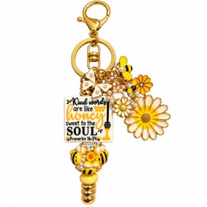 May include: A gold-toned keychain featuring a square charm with the text Kind words are like honey sweet to the SOUL Proverbs 16:24. The accessory includes charms of a white butterfly, yellow and black bees, and white daisies with yellow centers.