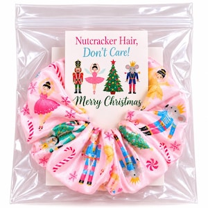 May include: A pink fabric hair scrunchie featuring a repeating pattern of nutcrackers, ballerinas, mice, Christmas trees, and candy canes. The scrunchie is packaged with a card that reads: Nutcracker Hair, Don't Care! Merry Christmas.