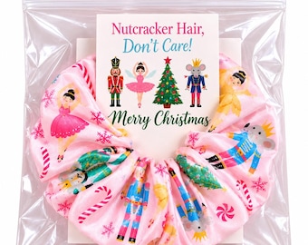 May include: A pink fabric hair scrunchie featuring a repeating pattern of nutcrackers, ballerinas, mice, Christmas trees, and candy canes. The scrunchie is packaged with a card that reads: Nutcracker Hair, Don't Care! Merry Christmas.